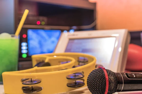 Black microphone in karaoke club, with remote controller, melon and strawberry soda drinks, yellow tambourine and screen for singing music on stage party.