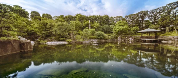 Central pond of Mejiro Garden which is surrounded by large flat stones under the foliage of the momiji maple trees and pines. The sky is reflected symetrically in the water of the pond. — Stock Image Central Pond Mejiro Garden Which Surrounded Large Flat Stones Foliage Royalty Free Stock Photos