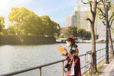 Japon kadın hakama kimonosu giymiş ve yüz maskesi takmış Tokyo İmparatorluk Sarayı 'nın hendek korkuluklarına dayanan bir Tokyo seyahat rehberi okuyor..