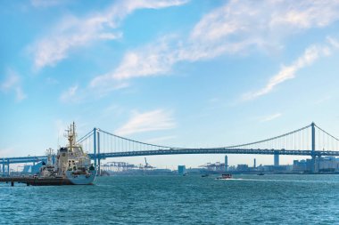 Tokyo Körfezi kıyılarına demirlemiş bir tekne. Gökkuşağı Köprüsü 'nün önünde. Arka planda Tokyo Limanı' nın Uluslararası Konteyner Terminali 'nin vinçleri var..