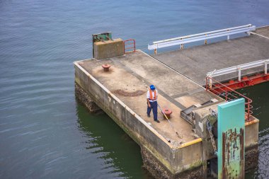 kanagawa, japan - july 19 2020: Dockworker wearing a life jacket using a boat hook to grab the hemp mooring line of the Kanayamaru ferry and hang it on a bollard of the pier of Kurihama port. clipart