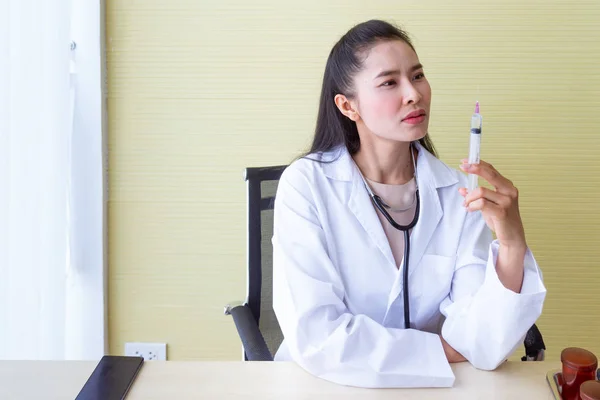 The female Asian doctor wearing a white robe sitting in a chair with a stethoscope on the neck be a serious face and preparing a syringe for patients.