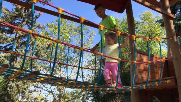 子供の つのクリップは 夏の晴れた日に遊び場でロープの吊り橋を渡る ビッグ ブラザーのティーンエイ ジャーは 妹の幼児をサポートします 兄弟が一緒に遊ぶ屋外 それ以上の年齢の少年と小さな女の子 — ストック動画