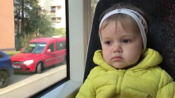 小女孩坐在运行的公共交通中 好奇地从窗户往外看 幼儿盯着电车窗外 城市里的小旅行者 孩子在里面坐公共汽车去 小乘客 — 图库视频影像
