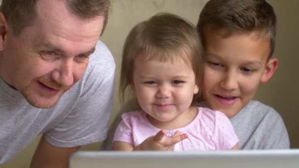 家长和两个孩子看着笔记本电脑屏幕一起玩在家里 儿子和女儿在看动画片笑 男人和兄弟姐妹 大哥和小妹妹希望展示和微笑 男孩和女孩在成人的控制下学习 — 图库视频影像