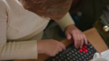 Kıdemli kadın iş yerinde klavyede yazıyor, üstte görüntü. Yaşlı kadın bilgisayarda çalışıyor, masada bardak. Büyükanne modern teknolojiyi kullanıyor, e-posta yazıyor. Bilgisayar, internet kullanan yaşlı iş kadını. Yaşlı, kapalı yaşam tarzı. Büyükanne masada, ellerde