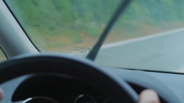 Araba sürerken arabanın ön camına yağmur yağıyor, yağmur algılayan silecekleri hareket ettiriyor, yakınlaşıyor. Ön ekranda ıslak yolda ulaşım sürücü. Odaklanmamış gösterge paneli, kontrol paneli, yağmurlu bir günde elleri ile direksiyon simidi. Yağmur saçılımı ile cam odaklanın