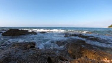 Deniz gelen dalgalar köpük yıkama kayalık kıyı, beyaz yelken uzak, martı mavi berrak gökyüzüne karşı su üzerinde uçan. Deniz köpüğü ile yaklaşan deniz dalgaları yıkanan taş kıyı, zemin sıçraması, kuş sineği. Kara saldalgası kayaları kırma