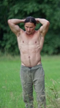 A fit man with long hair, A man ties his long hair into a ponytail before a workout in a green field, showcasing his impressive muscular physique in nature. Slow motion