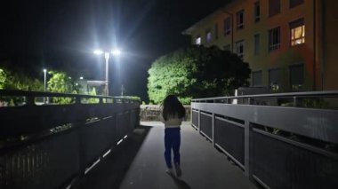 A young girl walks alone on a bridge that is illuminated by streetlights, surrounded by the nighttime cityscape