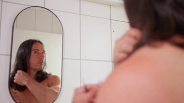 A man cuts his long black hair in front of a mirror. A tranquil, reflective moment that showcases the importance of selfcare and grooming in a bathroom setting. High quality 4k footage
