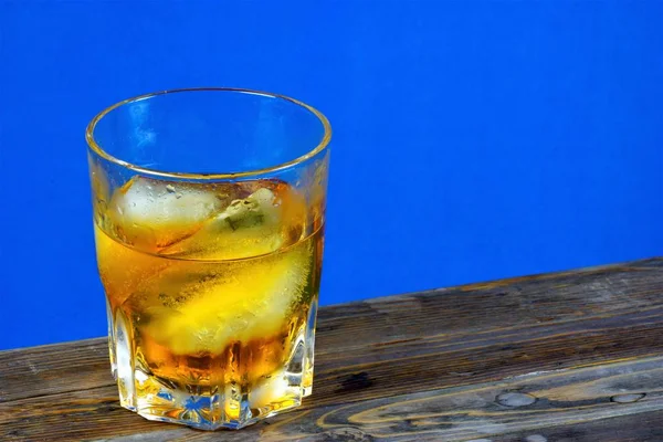 Alcoholic drink with ice on wooden table top, blue background. Strong aromatic alcoholic drink, from different types of grain - whiskey or Bourbon. Cognac or brandy - from grape must.