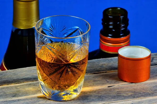 Alcoholic drink in a glass on a wooden table top, blue background. Strong aromatic alcoholic drink, from different types of grain - whiskey or Bourbon. Cognac or brandy - from grape must.