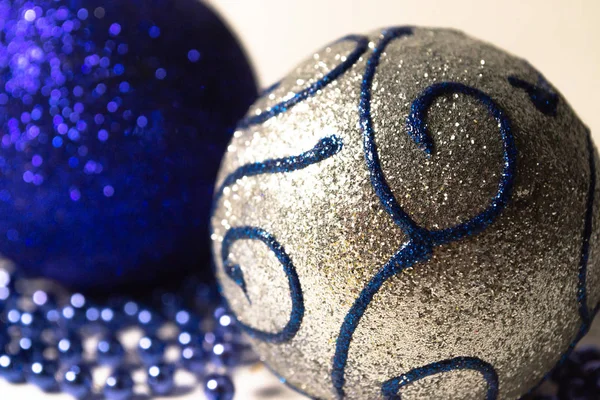 two Christmas tree toys shiny balls blue and silver with pattern lie on white background with blue beads closeup silver ball in foreground macrophotography