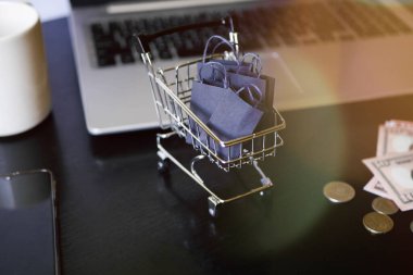 Çantalı alışveriş arabası dizüstü bilgisayarın yanında duruyor. Kara Cuma. Satış duyurusu. E-ticaret işi. İnternet mağazasından sipariş vermek. Güneş ışınları 