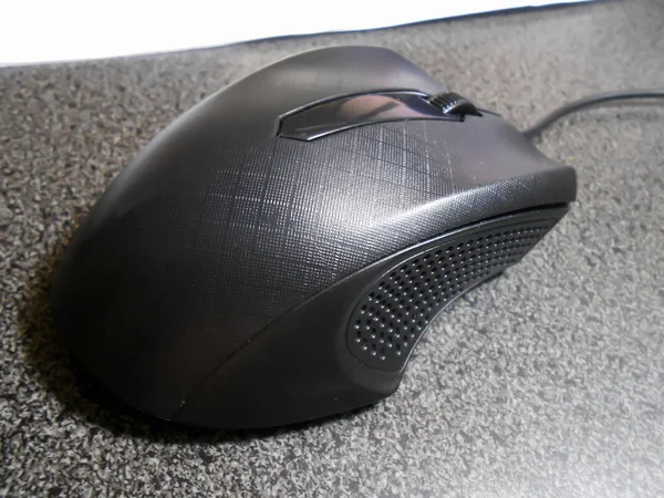 On a black spotted desk top and attached to a cable is a black computer mouse with lined patterns at the top of its body, and indent comfortable grit textured thumb and finger rest to the sides. To the top front is a scroll wheel button.