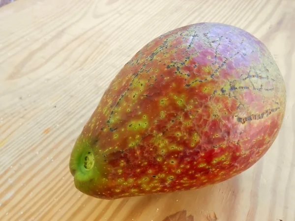 Ripe avocado pear with blemished, rough, marked, and spotted skin. The fruit is placed on the surface of light colored wooden chopping board with close parallel light brown grain lines running diagonally. Also on the board are spots and blemishes.