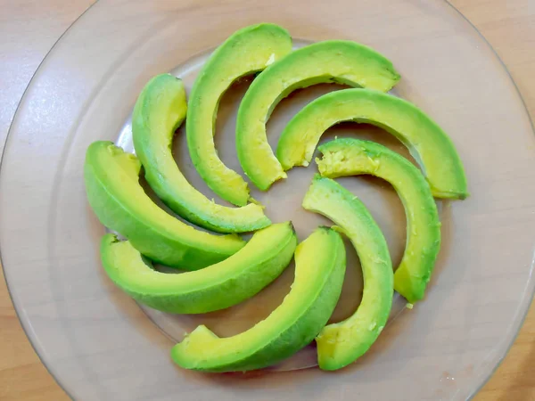 Indoor, on brown plastic laminate sheeting surface of natural wood texture, in transparent plate are slices of nutritious, uncooked, ripe avocado also known as alligator or butter pear, avocat, arranged to form swirling circle.