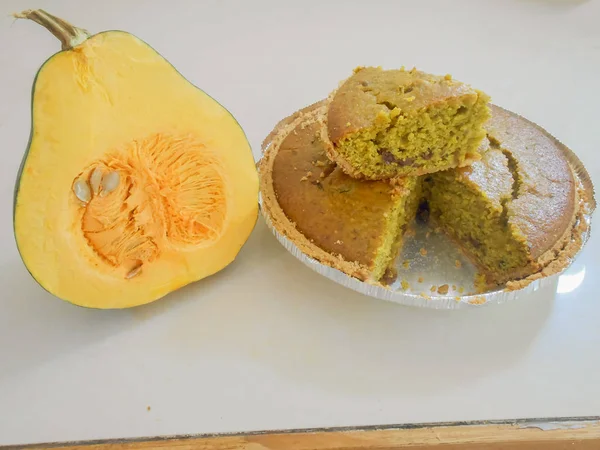 Cake with ingredients including nuts and raisons baked in a piecrust. A triangle gap of a thick slice cut out and placed on top showing the inside texture. Beside the aluminum containing the pastry is a cross section of a small pumpkin.