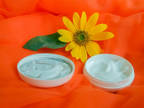 Colorful display of white skin cream on orange mesh material with small yellow sunflower, having a green leaf attached, in the background. The cosmetic is packaged in white container and left opened with inside of cover turning up.