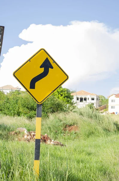 Yellow road sign pannel with black double bending arrow pointing up. The post of the sign is yellow and black. In the background are grass, trees, and building. The blue sky has white puff of cumulus cloud.