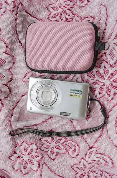 Small, silver point and shoot camera with black strop handle beside pink and black pouch on floral, pink and white lace fabric.
