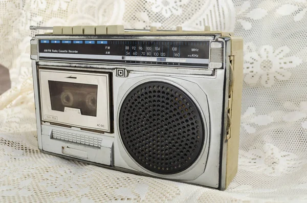 Old silver and black AM FM radio and cassette recorder with analog tuning wheel on ivory color floral laced fabric