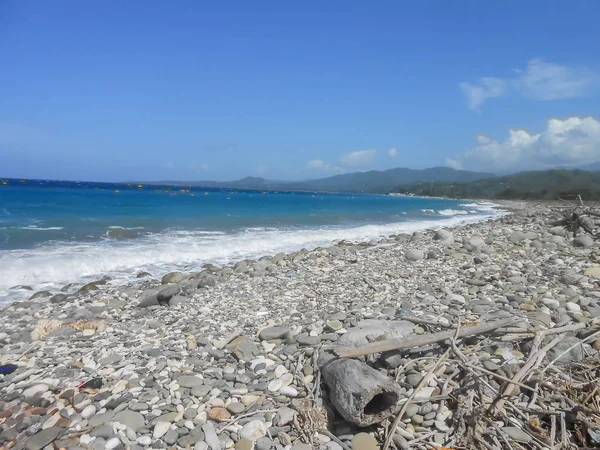 A fair sunny day at a lonely beach. Bright blue sky reflects in the ocean water as its wave rolls calmly to rocky shore.with debris of sticks, stones, and martar, In the distant sky is small patch of cumulus clouds.