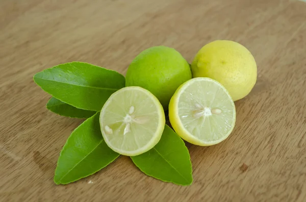 Two key limes ripen at different stages, and one cut in halves laid out on a leafy stem of it tree that is resting on a brown ply board surface. The lime cross section shows it radial segments of juice sacs, and seeds in its radial segments.