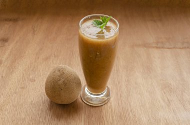 Bir tahta masa yüzeyinde yeşil nane ile süslenmiş kahverengi sapodilla smoothie ile dolu berrak bir bardak vardır. Bardağın yanında, adı içki olan olgun bir meyve var..