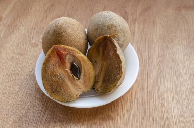 Bir tahta yüzeyinde, sapodilla ya da naseberry olarak da bilinen manilkara zapota ile dolu küçük beyaz bir tabak bulunur. Ön tarafın enine kesitinde düz bir sırt tohumu var..