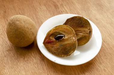 Kahverengi sapodilla kesitleri beyaz bir tabakta tohum ile kahverengi doğrusal tahıl tahıl yüzeyinde. Meyvenin yarısına küçük siyah bir tohum iliştirilmiş. Tabağın yanında koca bir meyve var..