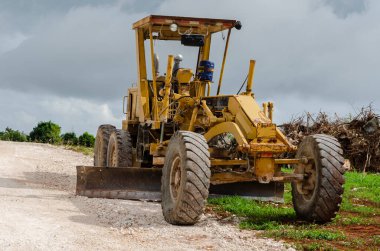 Bulutlu gökyüzünün altında bıçağı yerde dinlenen bir motorlu greyder, bitmemiş bir yolun kenarına park edilmiş, bir ön tekerleği çimenli zeminde, diğeri ise marl yüzeyinde..