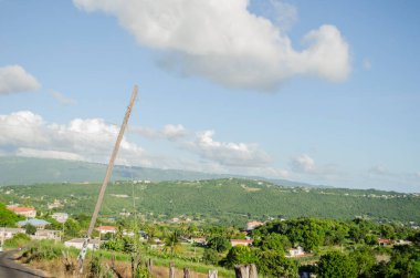 Gün parlak ve güneşli. Mavi gökyüzünde ilerleyen yoğun bulutlara ve ufuktaki uzak dağdan yakınlardaki yerleşim bölgesine uzanan yeşil yemyeşil bir bitki örtüsünün üzerinde asılı olan bulutlara rağmen..