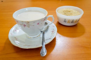 Formika yüzeyinin üstünde, fincanı sıcak çikolatalı içecek ve çay kaşığıyla dolu beyaz bir çiçek çayı seti var. Yanında yumurtadan yapılmış muhallebi dolu küçük bir tabak var..