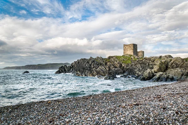 This is an Irish castle on Dough Island, Donegal, Ireland.  The castle is purched on rocks that jut out into the sea. in the forground is a stone beach.
