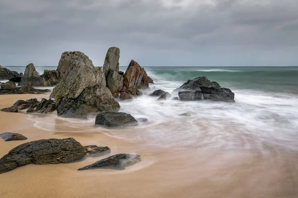 Bulanık dalgalar kayaların İrlanda kumlu bir plaj üzerinde çevresinde swirling. Gökyüzü fırtınalı ama bulanık dalgalar Kayaların etrafında yumuşak bir ışık oluşturmak.