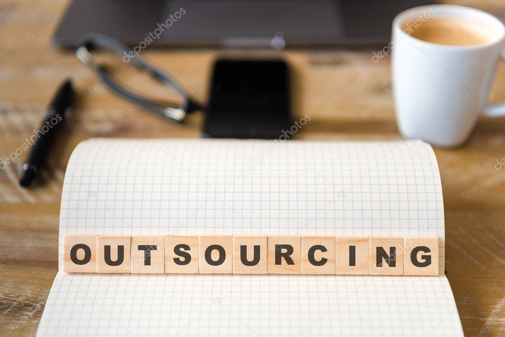 Closeup on notebook over wood table background, focus on wooden blocks with letters making Outsourcing text. Concept image. Laptop, glasses, pen and mobile phone in defocused background