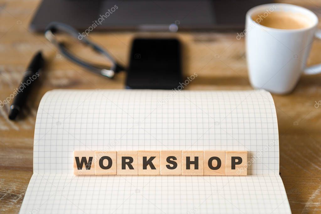 Closeup on notebook over wood table background, focus on wooden blocks with letters making Workshop text. Concept image. Laptop, glasses, pen and mobile phone in defocused background