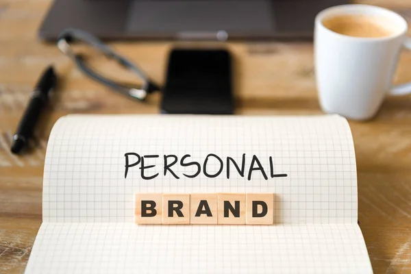 Closeup on notebook over vintage desk surface, front focus on wooden blocks with letters making Personal Brand text. Business concept image with office tools and coffee cup in background