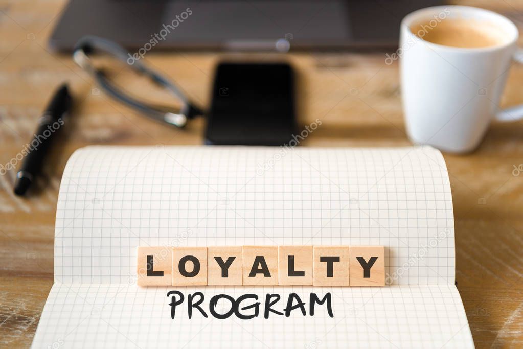 Closeup on notebook over vintage desk surface, front focus on wooden blocks with letters making Loyalty Program text. Business concept image with office tools and coffee cup in background