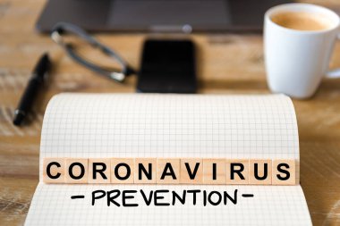 Ahşap masa arkasındaki deftere yakın çekim, Coronavirus Prevention yazımı yapan harflerle ahşap bloklara odaklan. Bilgisayarlı ve cep telefonlu iş kavramı çözülmüş bir arkaplanda