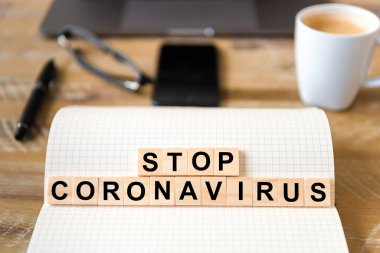 Ahşap masa arkasındaki deftere yakın çekim, Coronavirus 'u durduran harflerle ahşap bloklara odaklan. Bilgisayarlı ve cep telefonlu iş kavramı çözülmüş bir arkaplanda