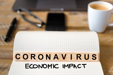 Ahşap masa arkasındaki deftere yakın çekim, Coronavirus Ekonomik Etki yazımı yapan harflerle ahşap bloklara odaklan. Bilgisayarlı ve cep telefonlu iş kavramı çözülmüş bir arkaplanda