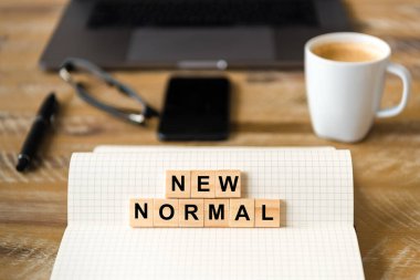 Not defterini ahşap masa arkaplanına yakın tut, New Normal 'ı yazı haline getiren harflerle tahta bloklara odaklan. Bilgisayarlı ve cep telefonlu iş kavramı çözülmüş bir arkaplanda