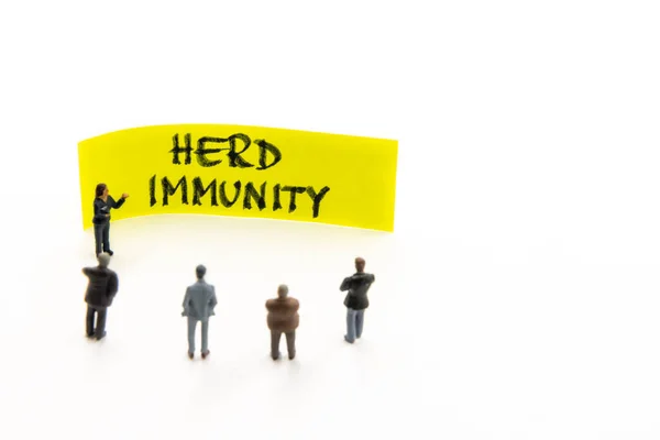 Meeting with miniature figurines posed as business people standing around post-it note with Herd Immunity handwritten message in background, minimalist abstract concept with focus on text