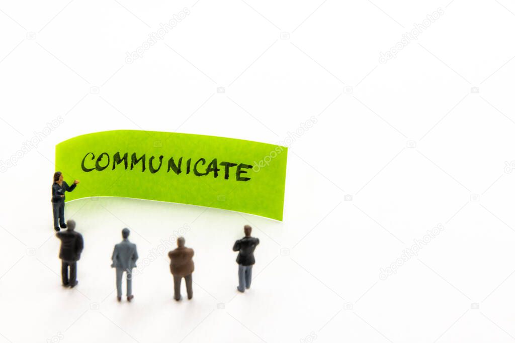 Meeting with miniature figurines posed as business people standing around post-it note with Communicate handwritten message in background, minimalist abstract concept with focus on text