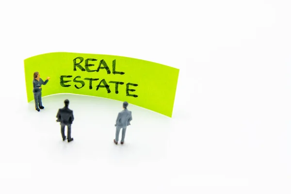 Presentation meeting with miniature figurines posed as business people standing in front of post-it note with Real Estate handwritten message in background, copy space on the right