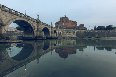 Gün batımında Sant Angelo Kalesi'nin yan manzarasına sahip Roma'nın tarihi merkezinde Tiber nehri üzerinde Aurelius köprüsü. Sudaki taş köprünün yansımaları.