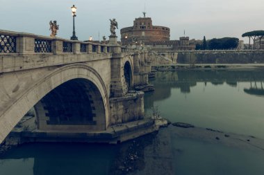 Gün batımında Sant Angelo Kalesi'nin yan manzarasına sahip Roma'nın tarihi merkezinde Tiber nehri üzerinde Aurelius köprüsü. Sudaki taş köprünün tarihi aydınlatması ve yansımaları